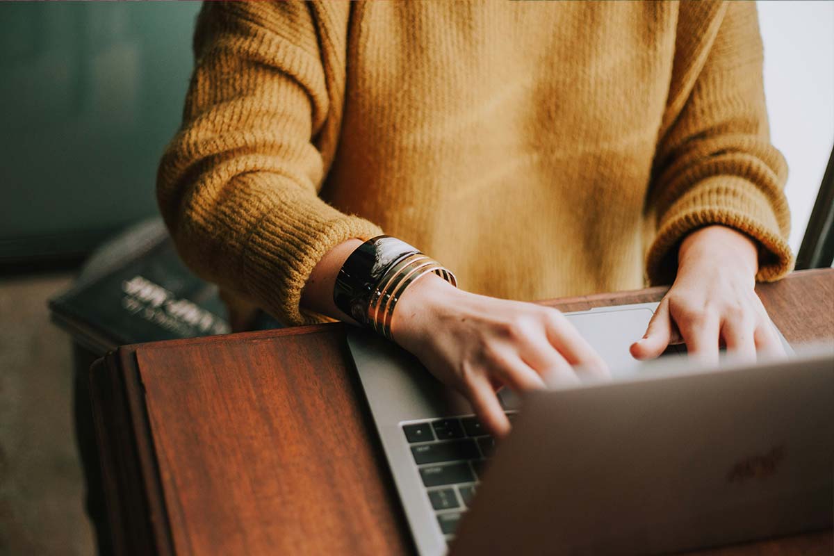 A person in a mustard sweater typing on a laptop at a wooden desk